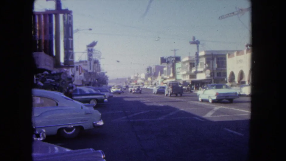 Wide 1962 Tijuana street view with classic cars, storefronts, commercial signs, utility poles, and traffic under a clear sky.