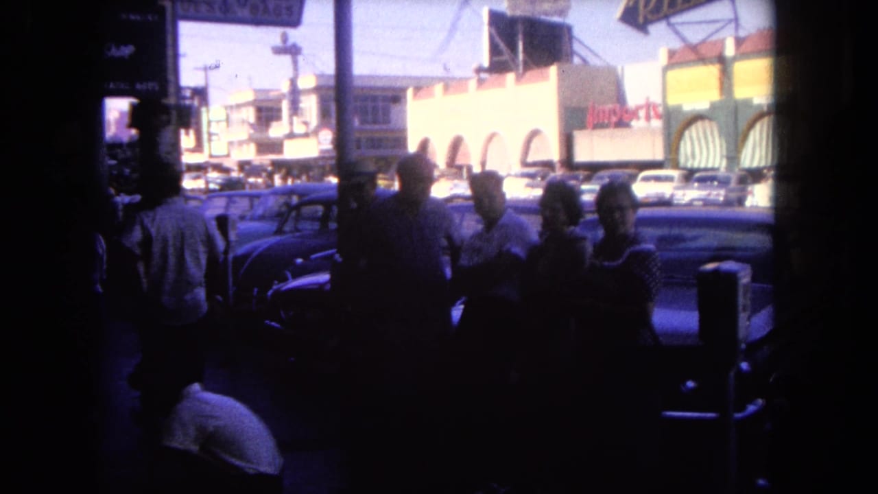 Pedestrians stand in shadow near parked cars, with storefronts and an Imports sign across the street.