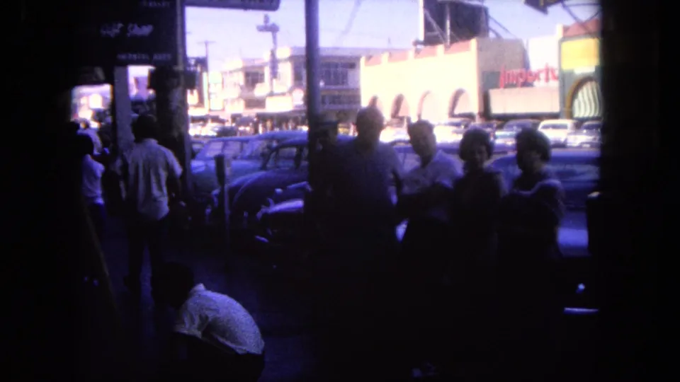 A shadowed group stands beside parked cars on a 1962 Tijuana commercial street, with storefront signage across the road.