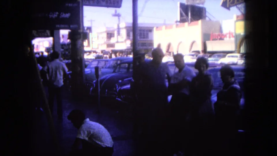 Late-frame 1962 Tijuana street view showing pedestrians in shadow, a crouching person, parked cars, and commercial storefronts.