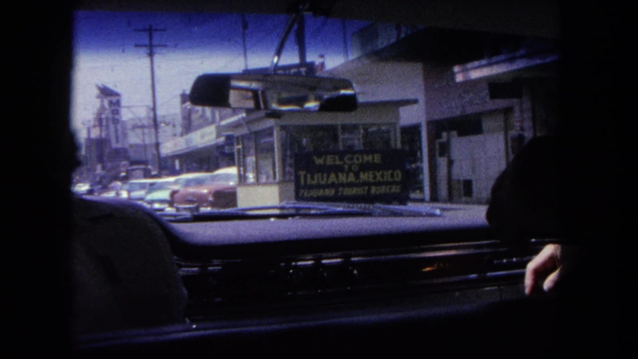 1962 Tijuana street seen from inside a car, with dashboard, rearview mirror, classic cars and a welcome sign visible ahead.
