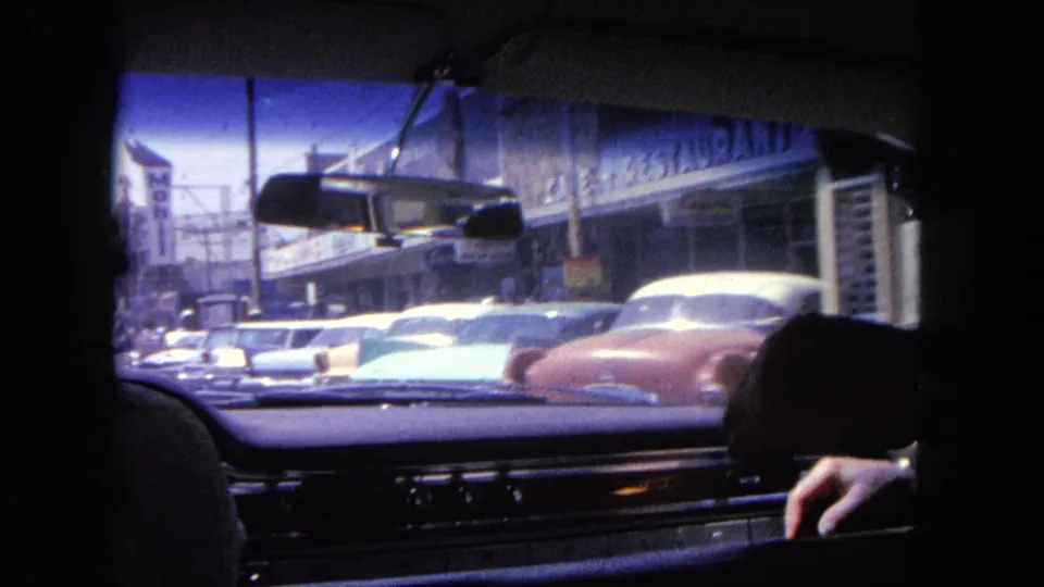 In-car 8mm view of downtown Tijuana traffic, with a silhouetted passenger, dashboard, mirror, storefronts and mid-century cars ahead.