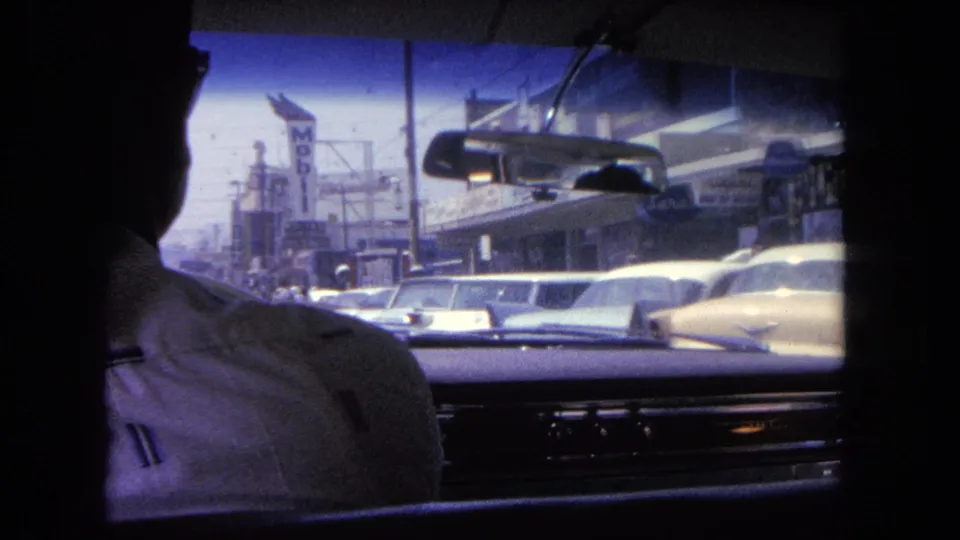 A 1962 in-car view down a Tijuana street, showing dashboard trim, rearview mirror, traffic, storefronts and a vertical motel sign.