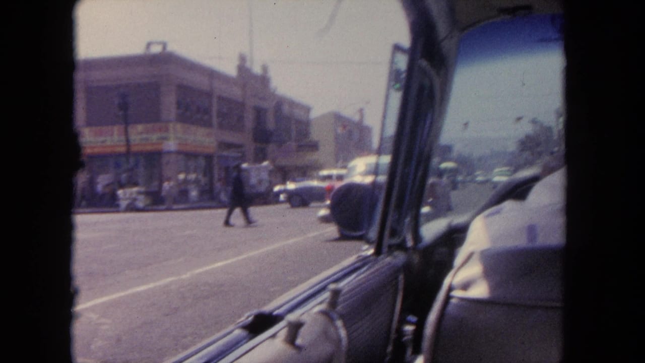 Street corner seen through a car window with pedestrians and classic cars in traffic.