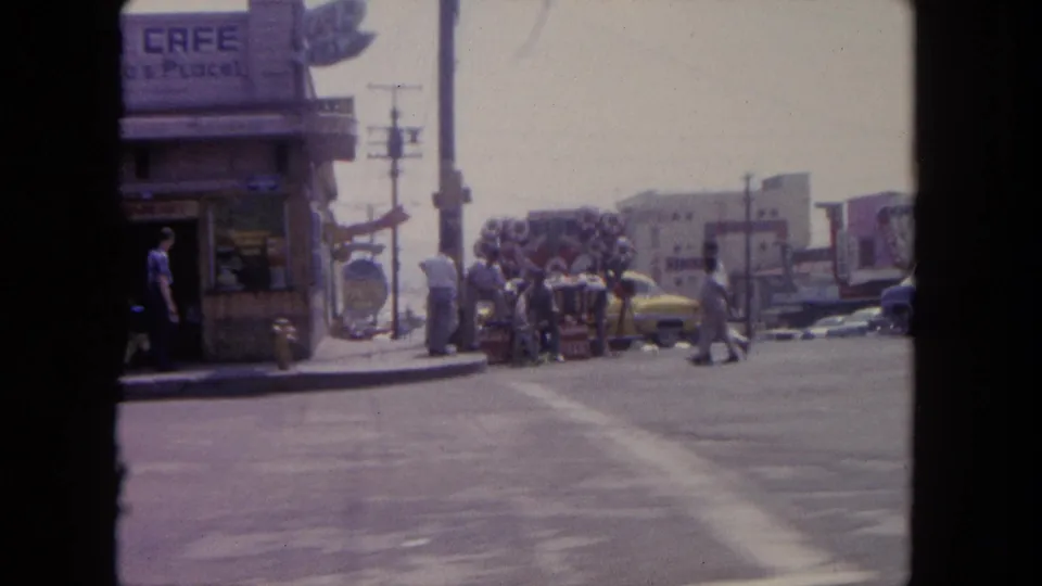 Tijuana corner with a cafe sign, sidewalk pedestrians, a yellow car, and utility poles in 1962 footage.