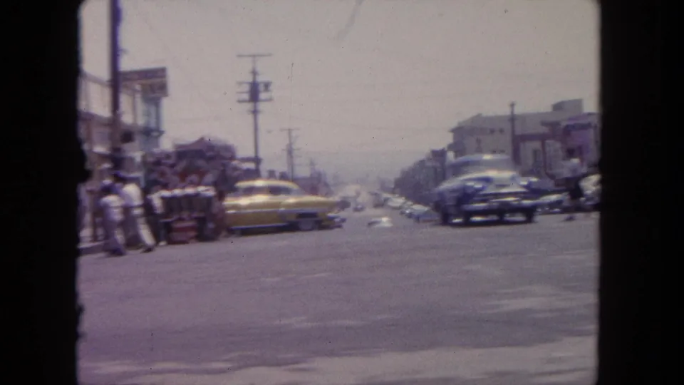 1962 Tijuana street scene with a yellow sedan, curbside pedestrians, utility poles, and passing cars.