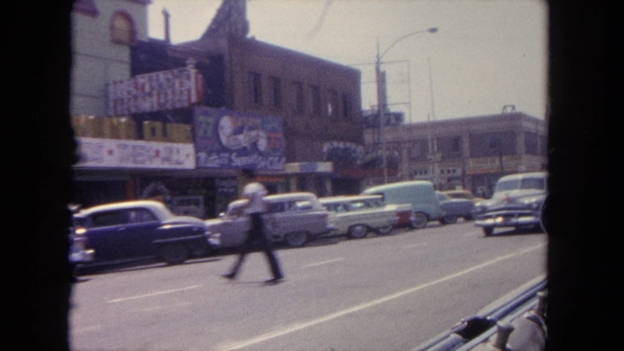 Street view with classic cars, pedestrians, storefronts, and a moving-car window edge.