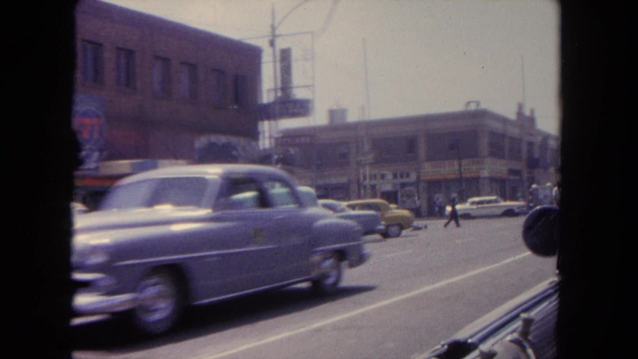 Moving-car view in Tijuana with a blurred early-1960s sedan, parked cars, storefronts, and pedestrians farther down the street.