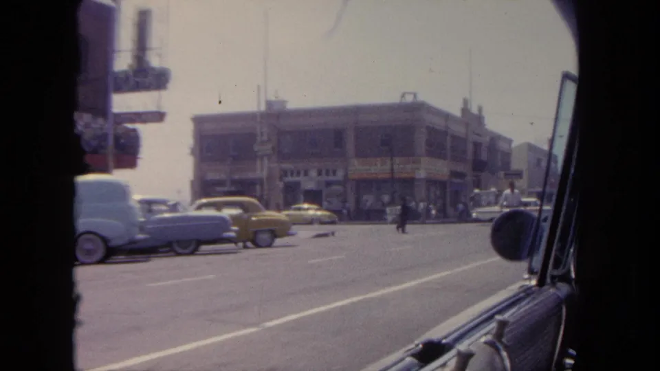 Tijuana commercial street seen through a car window, with parked mid-century cars, a yellow vehicle, storefronts, and pedestrians.