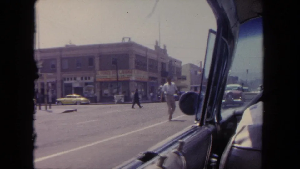 1962 Tijuana street scene with pedestrians crossing, classic cars, storefront signs, and the filming car's window and door frame.