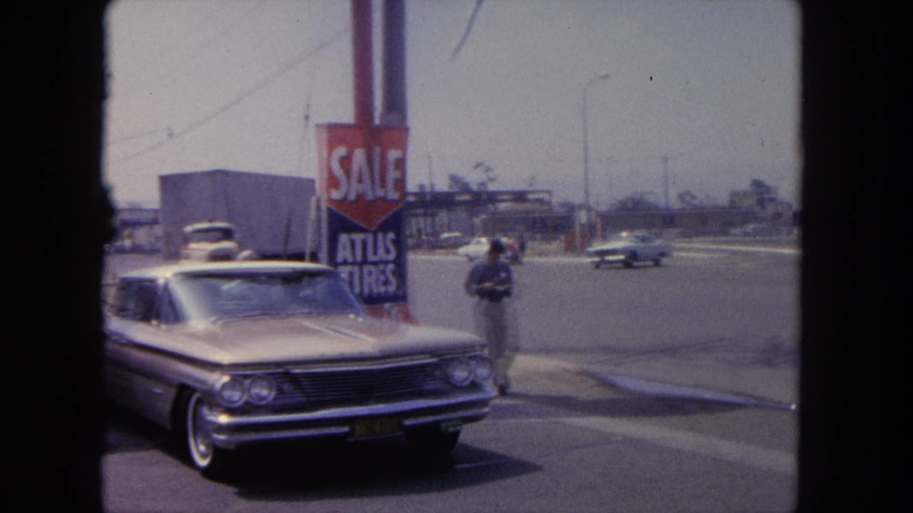 1962 Tijuana street view with a silver early-1960s sedan, Atlas Tires sale sign, traffic, and a pedestrian near the curb.