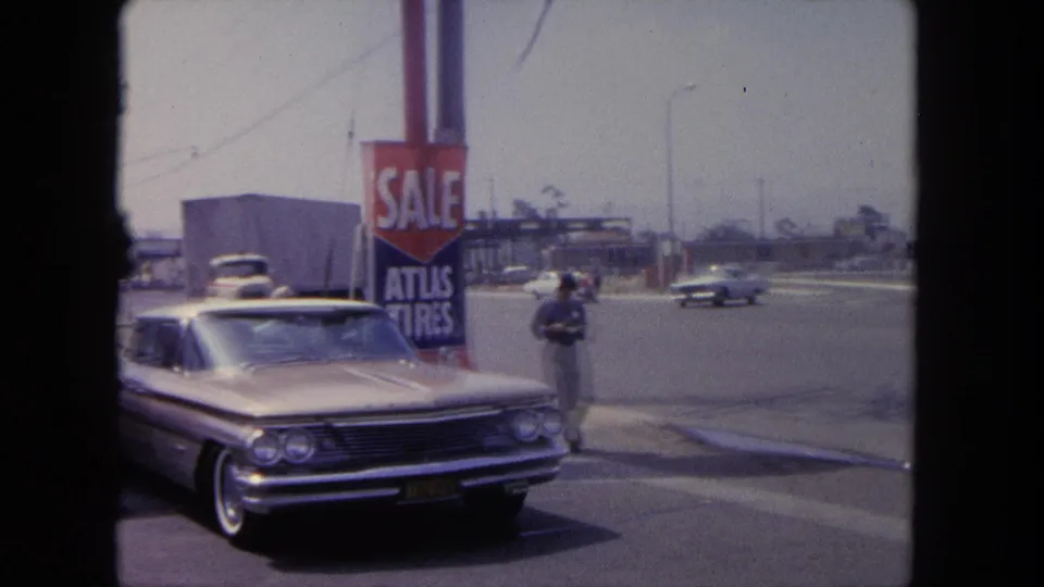 1962 Tijuana street view with a silver early-1960s sedan, Atlas Tires sale sign, traffic, and a pedestrian near the curb.