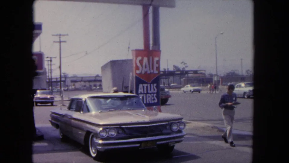 1962 Tijuana street scene with a parked sedan, prominent Atlas Tires sale sign, moving traffic, and a man walking at the right.