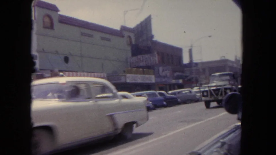 1962 Tijuana commercial street with vintage cars, storefront facades, parked vehicles, and roadway traffic filmed from a moving vehicle.