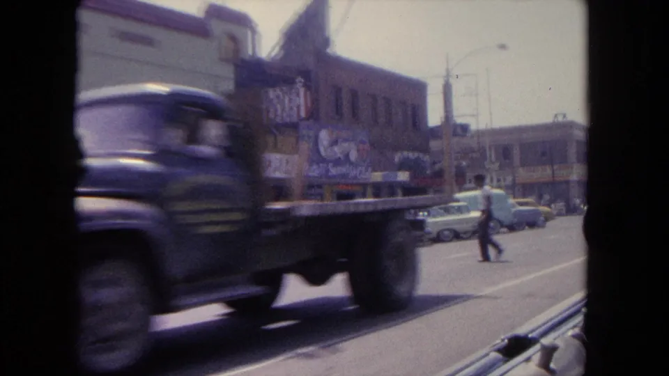 1962 Tijuana street view with a passing flatbed truck, storefront signage, pedestrians, parked cars, and sunlit roadway.