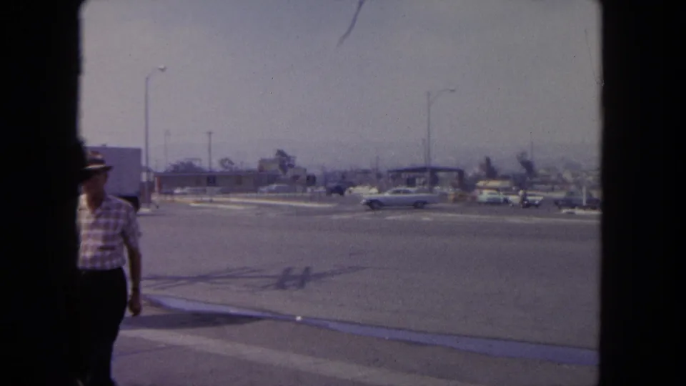 1962 Tijuana street view with early-1960s cars, low roadside buildings, streetlights, and a partly visible adult pedestrian near the frame edge.