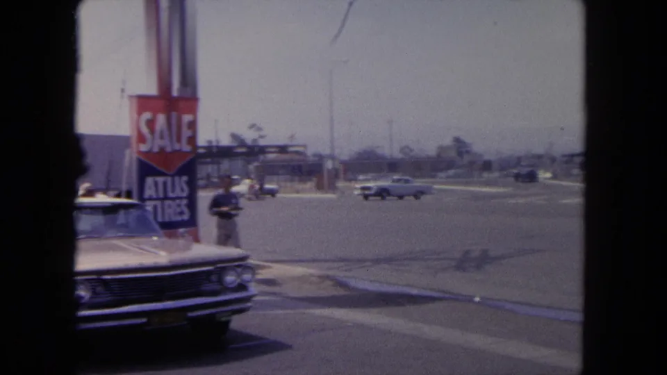 1962 Tijuana roadside view with a parked classic sedan beside a visible sale and tire sign, plus moving cars across the street.