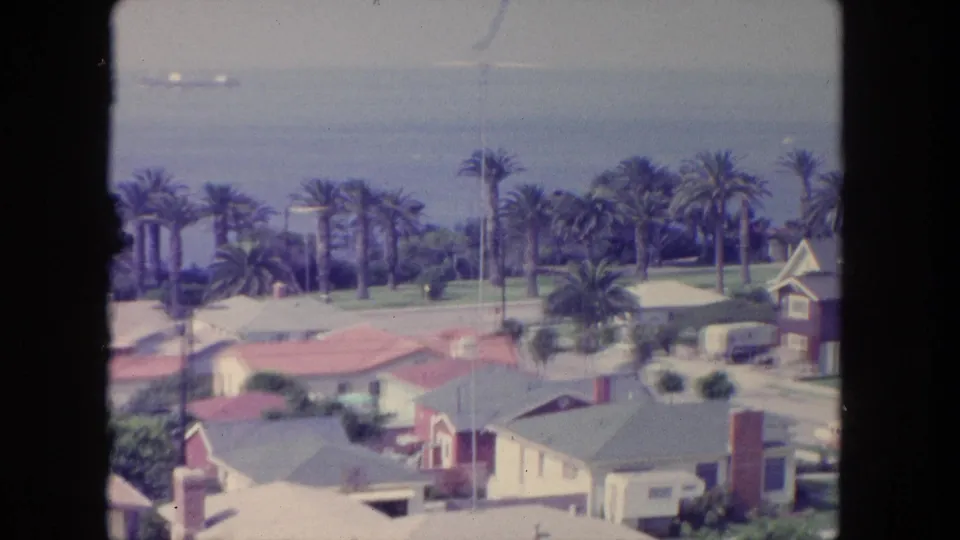 Soft 8mm frame of a 1962 Tijuana coastal neighborhood, showing rooftops, palms, ocean water, and a pale hazy sky.