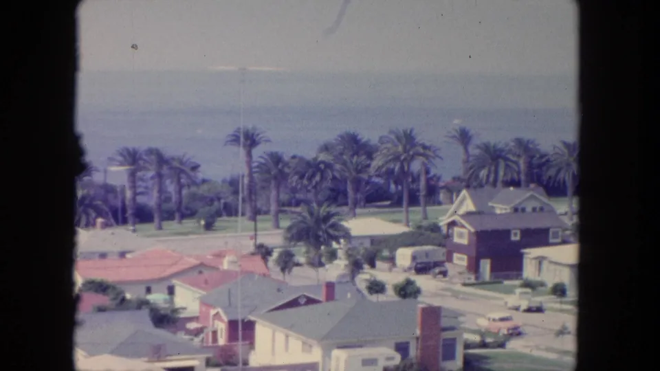 Elevated 1962 Tijuana view with coastal homes, a street edge, tall palms, ocean water, and a distant hazy horizon.