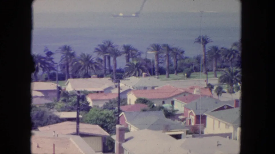 Elevated 1962 Tijuana view over red and pale rooftops, palm trees, and distant ocean under hazy light.