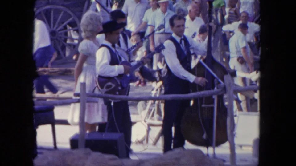 In 1962 Tijuana, two men perform with guitar and upright bass beside a woman in a white dress and a crowd behind a rail.