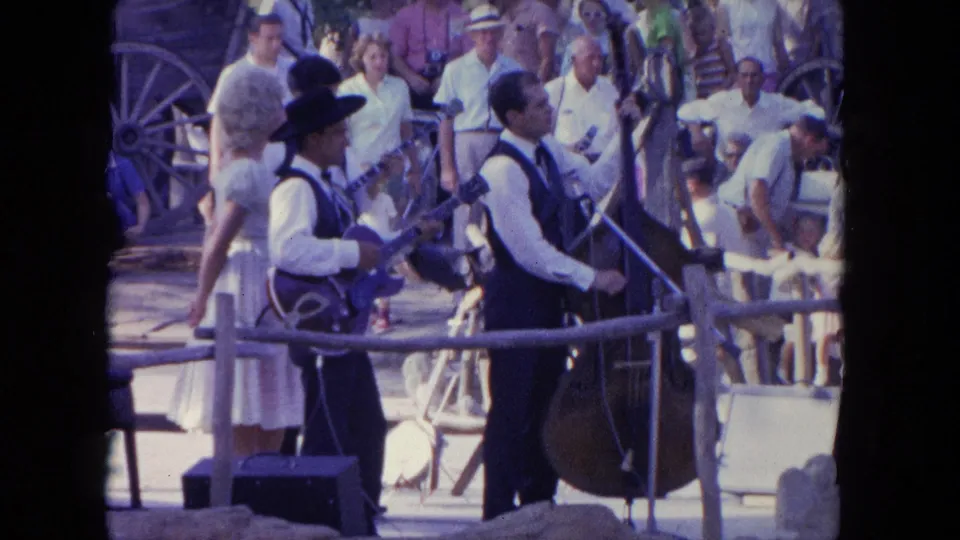 The Tijuana outdoor stage view shows the guitarist, upright bass player, nearby woman, and spectators in bright daylight.