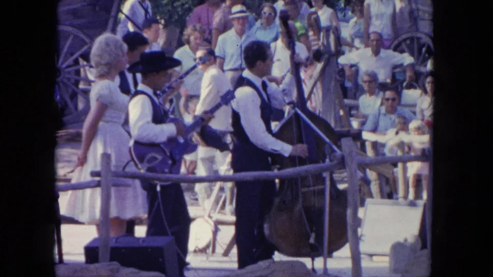 The 1962 Tijuana footage holds on the guitar and bass performers with a woman nearby and a crowd watching in sunlight.
