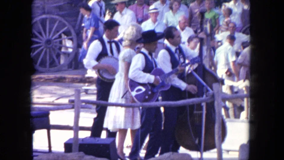 In the 1962 Tijuana scene, the camera frames musicians in western-style clothing with spectators and wagon wheel decor behind them.