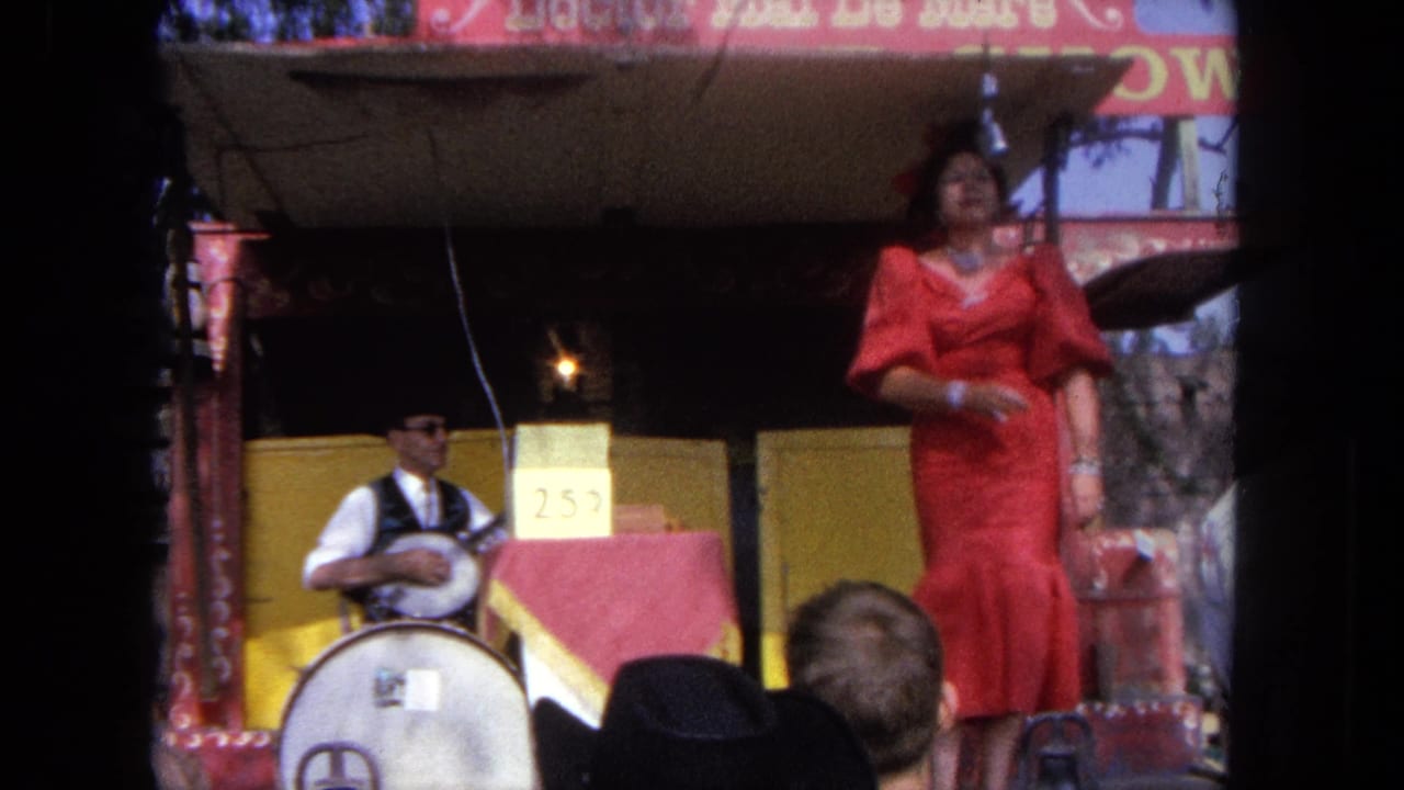 A woman in a red dress gestures mid-song on a Tijuana carnival stage while the seated banjo player performs at left.