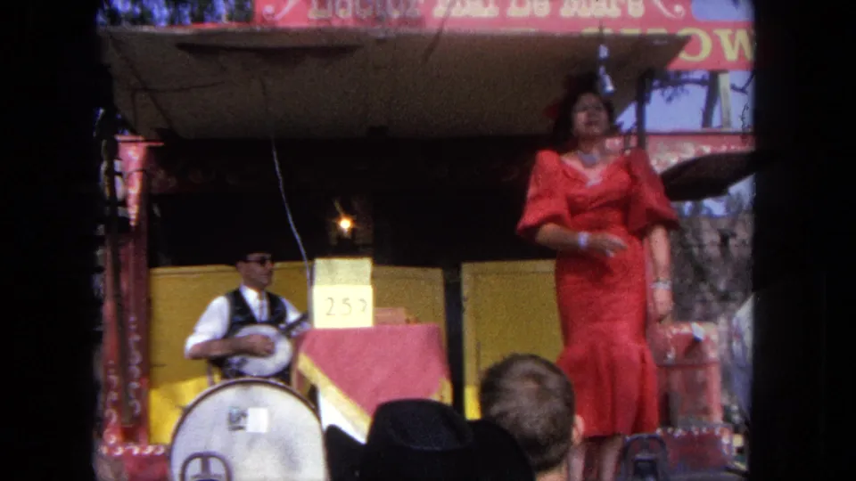 A woman in a red dress gestures mid-song on a Tijuana carnival stage while the seated banjo player performs at left.