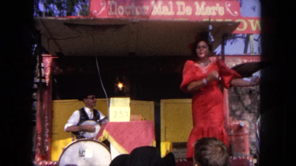 The Tijuana stage performer gestures toward the audience while a musician sits to one side with a drum and painted signage behind them.