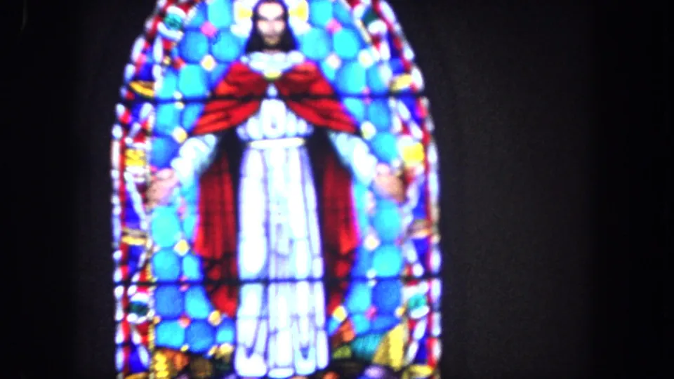 1962 Tijuana home-movie frame showing a stained-glass depiction of Jesus with a red cloak and white robe in a dark church interior.