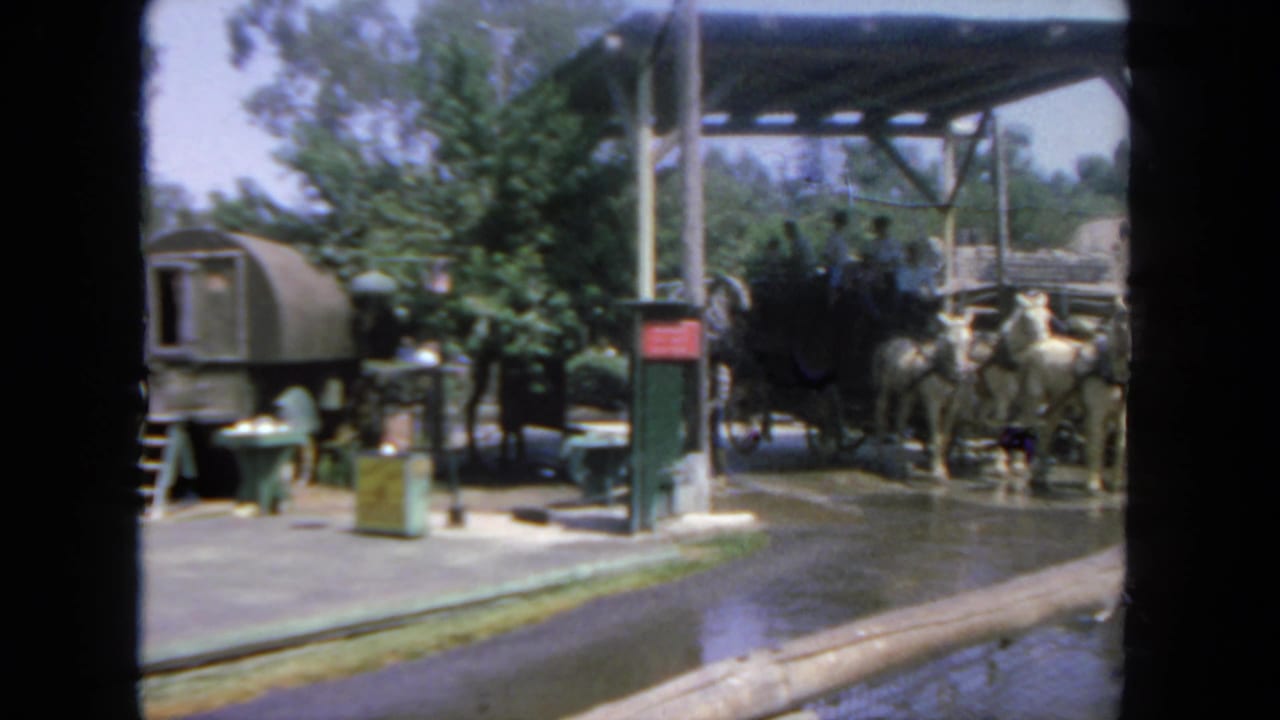 1962 San Diego frame with a covered hitching area, wet pavement, white horses, and a black stagecoach partly visible.