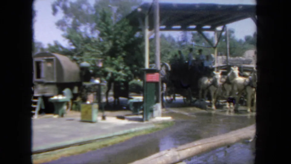 1962 San Diego frame with a covered hitching area, wet pavement, white horses, and a black stagecoach partly visible.