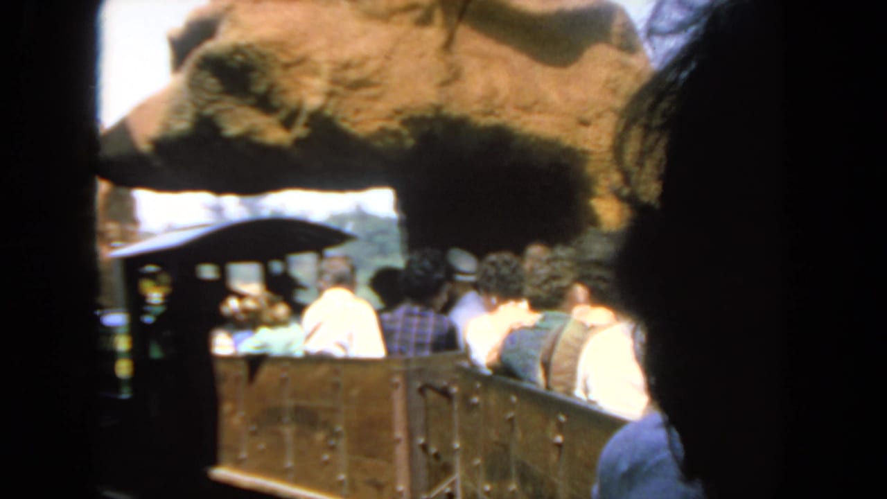 1962 San Diego home-movie frame showing seated passengers in open tour train cars moving through a rocky attraction setting.