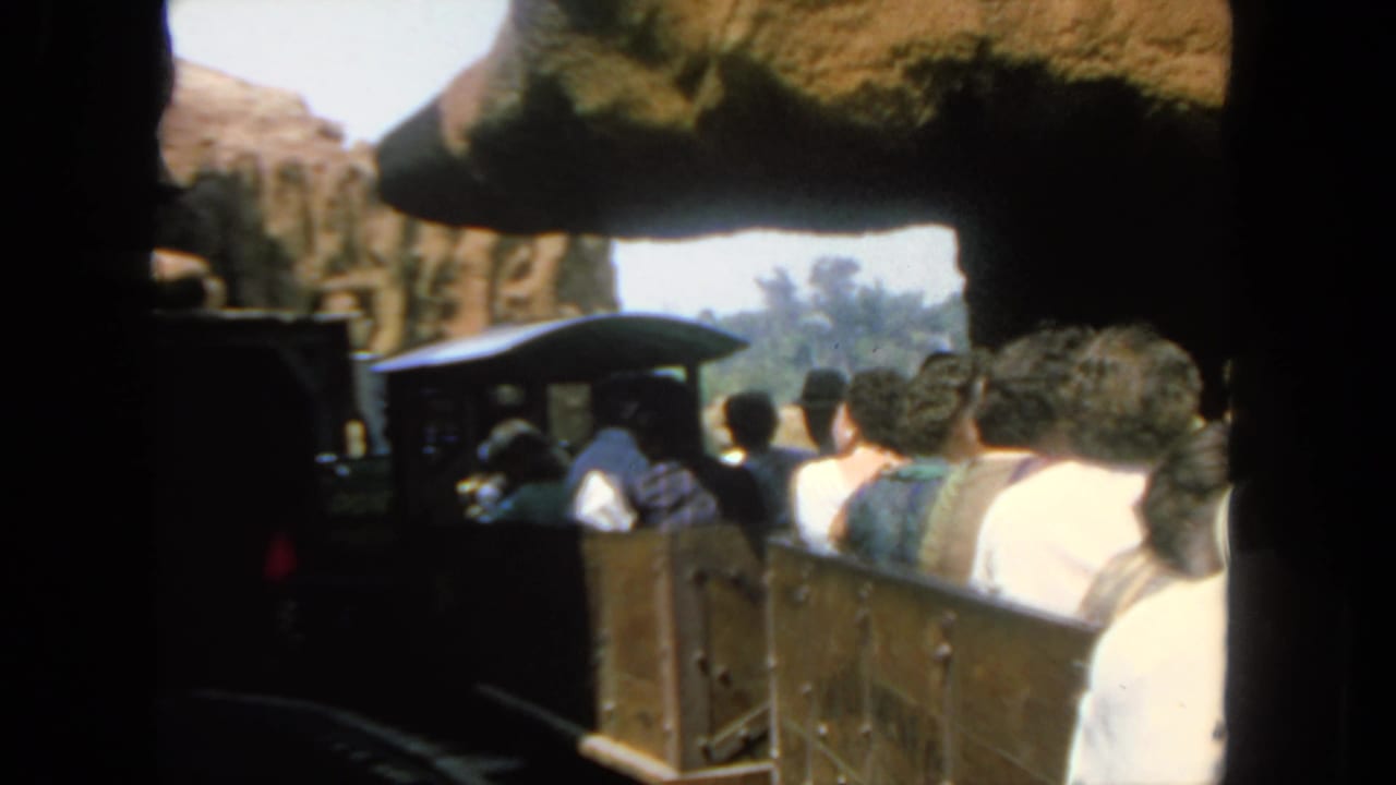 Visitors in casual 1960s clothing ride open tour train cars through a San Diego rocky attraction setting.