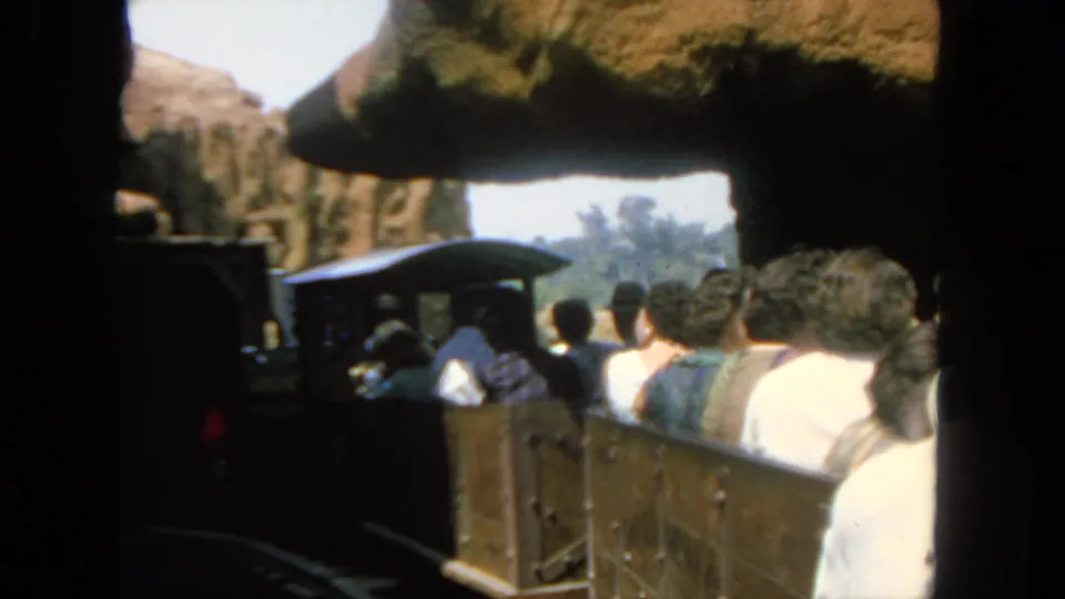 Visitors in casual 1960s clothing ride open tour train cars through a San Diego rocky attraction setting.