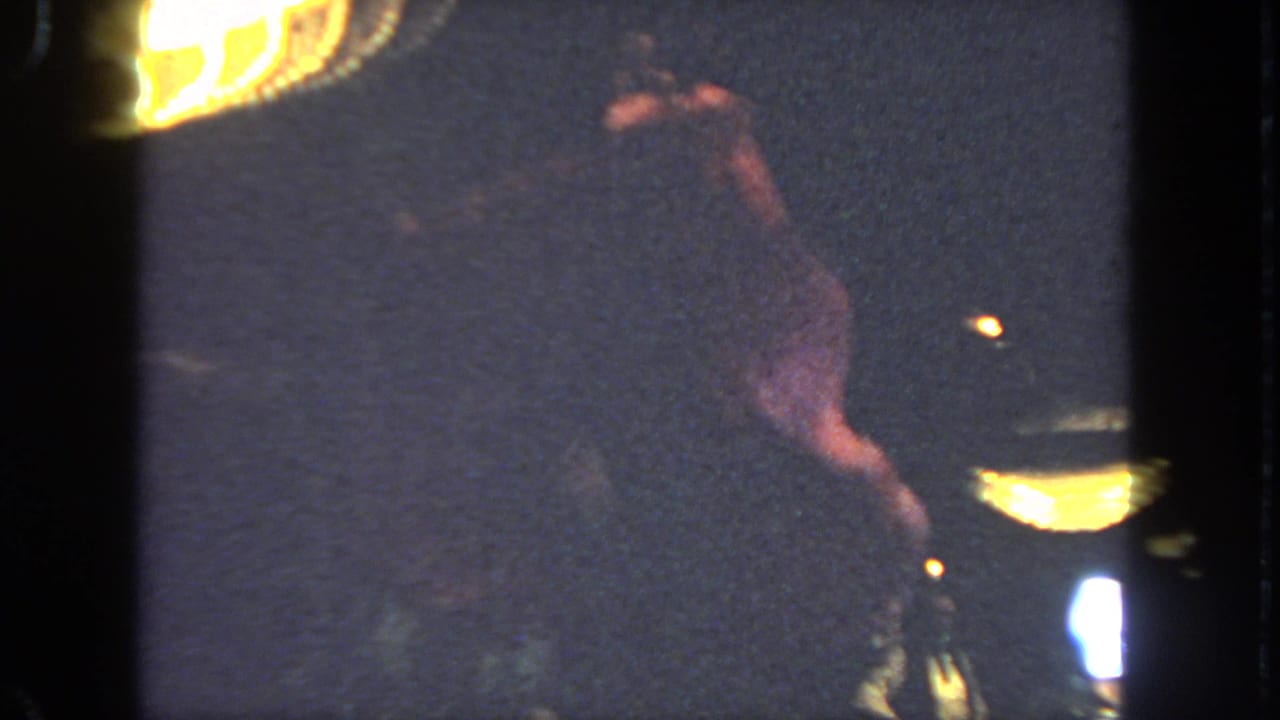 Red-dressed dancer bends or turns on a dim stage beside bright stage lighting.