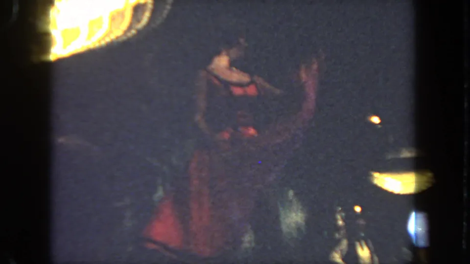 A red-costumed dancer raises one leg in a cancan pose on a dim stage, seen in grainy 1962 San Diego 8mm footage.