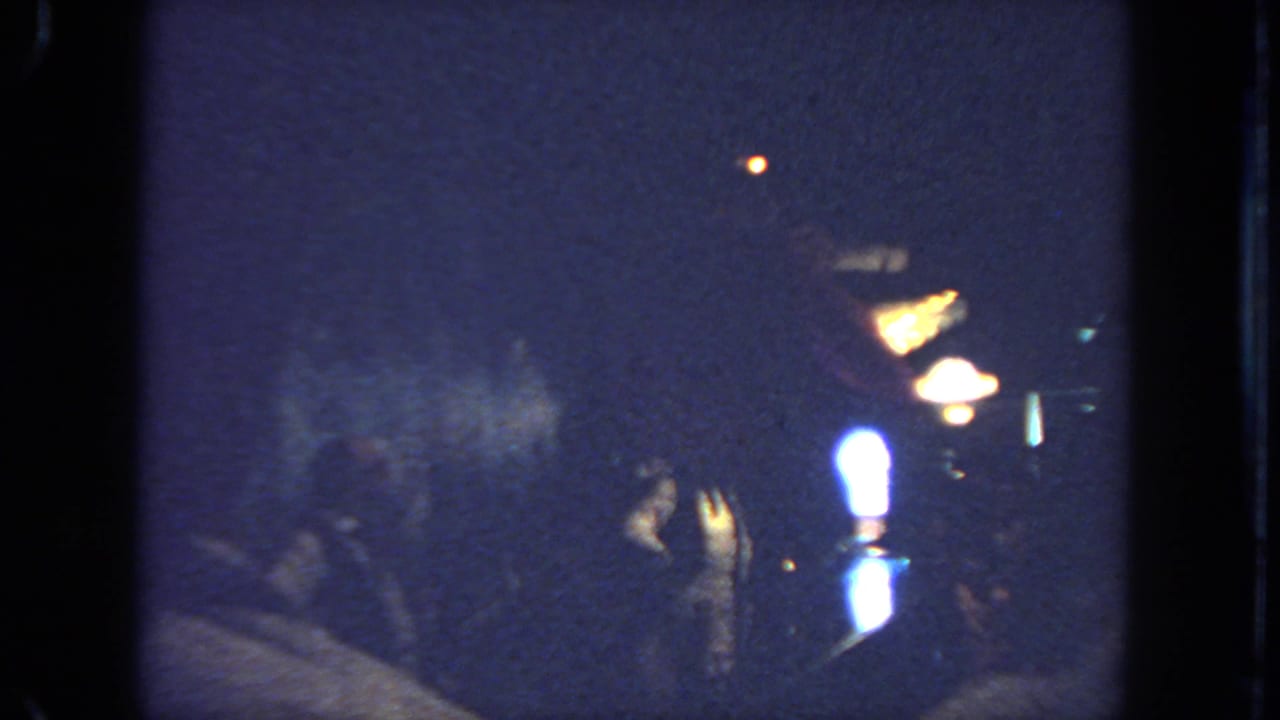 A dim indoor scene with a costumed dancer near warm lamps and shadowed spectators.