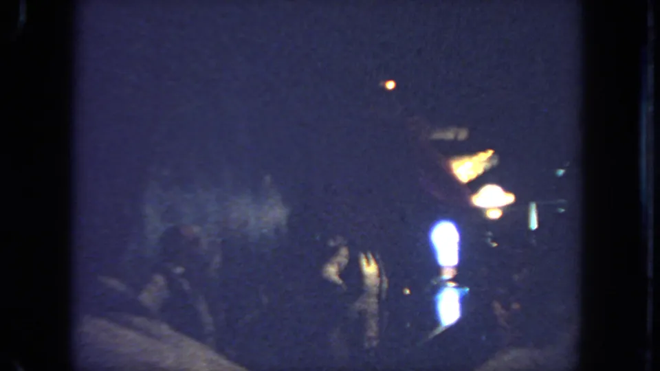 1962 San Diego 8mm frame showing a western-style dancer in low light, with warm lamps and people partly visible in the shadows.