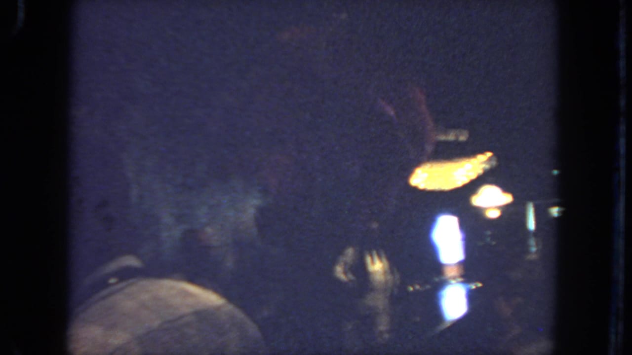 1962 San Diego home-movie frame with the dancer partly obscured as warm lamps glow over a dim indoor gathering.