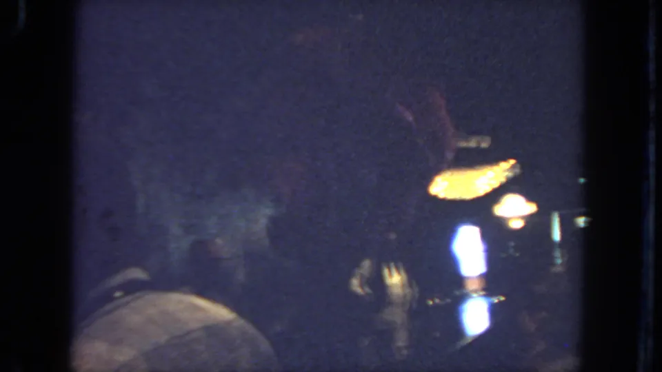 1962 San Diego home-movie frame with the dancer partly obscured as warm lamps glow over a dim indoor gathering.