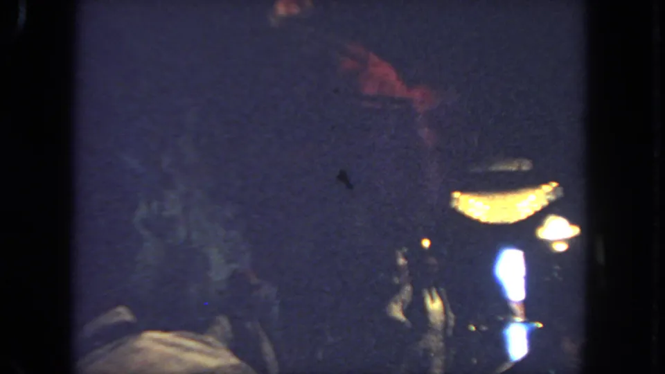 1962 San Diego 8mm frame showing the dancer's light skirt and upper body near warm lamps, with viewers barely visible in darkness.
