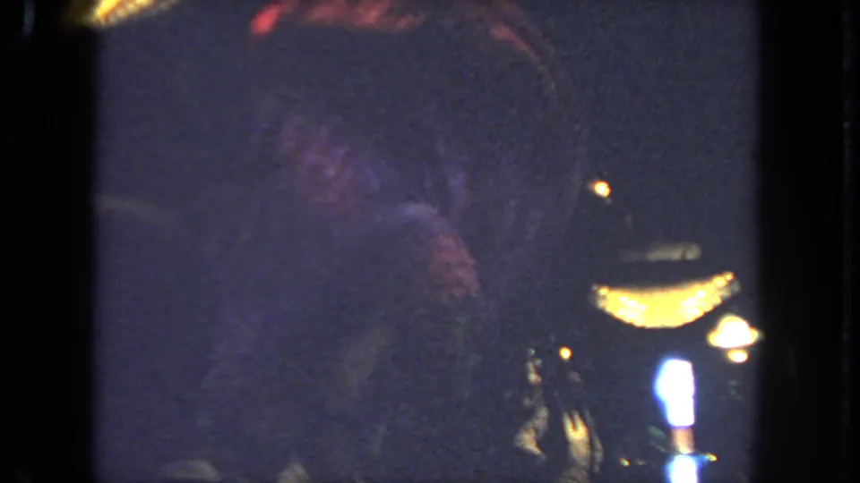 1962 San Diego home-movie frame with the dancer mostly in shadow beside warm lamps and indistinct spectators.