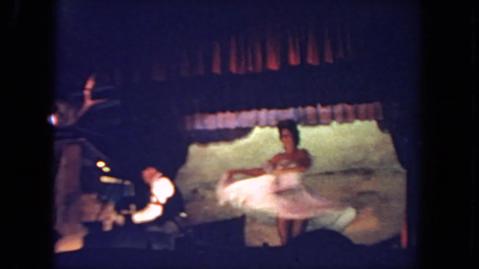 In this 1962 San Diego 8mm frame, a woman in a white dress spins on a dimly lit stage while a keyboard player sits to the side.
