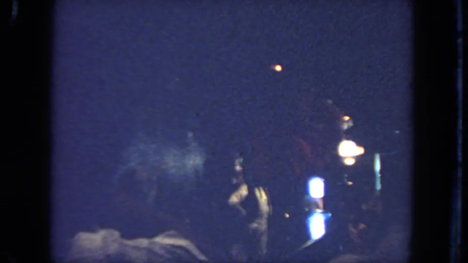 The home-movie frame drops into very low light, showing bright lamps, dark stage space, and faint figures or equipment near the bottom edge.
