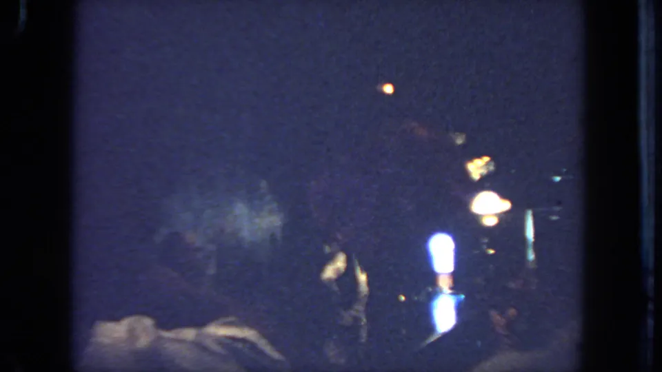 A dim 1962 San Diego 8mm frame shows bright stage lamps, blue-toned darkness, and indistinct figures or equipment in the performance area.