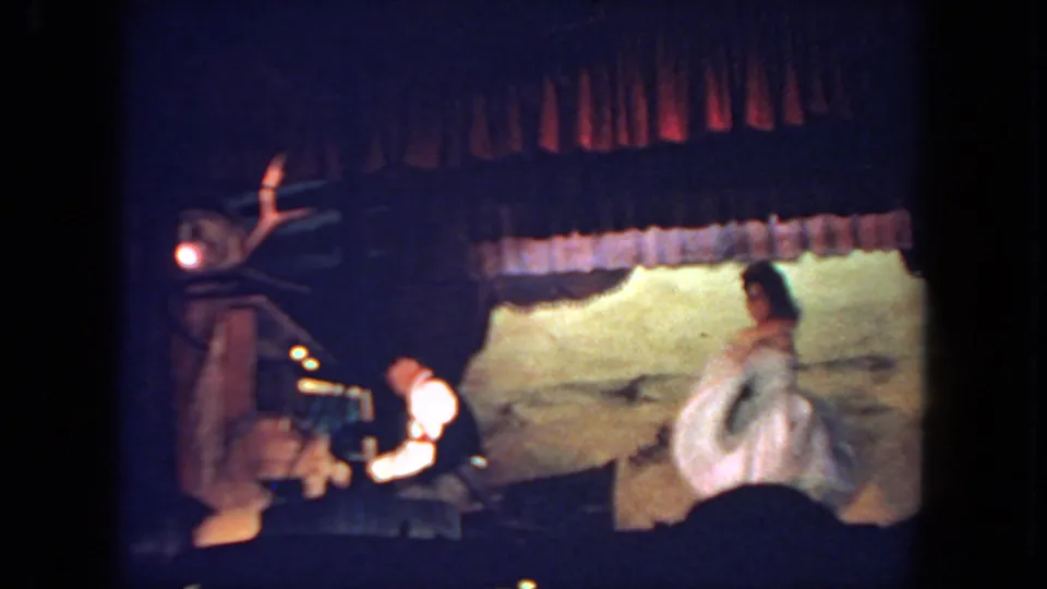 A handheld audience view shows the white-costumed dancer moving across the stage beside the piano accompanist, with curtains and a lit backdrop behind them.