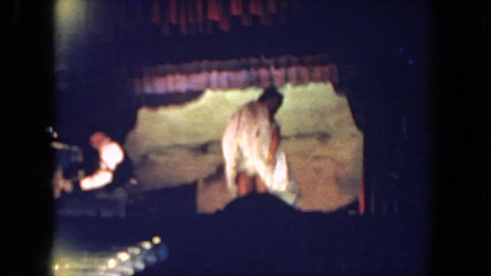 The dancer faces toward the backdrop in a white costume as the pianist is partly obscured at stage left, with audience silhouettes framing the lower edge.